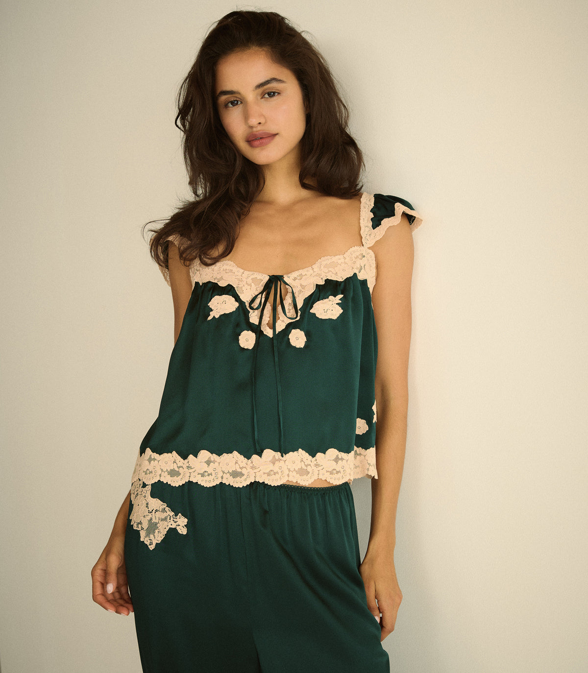 A woman wearing a dark green satin set with beige lace trim and a tie-neck detail, posed against a plain off-white background.