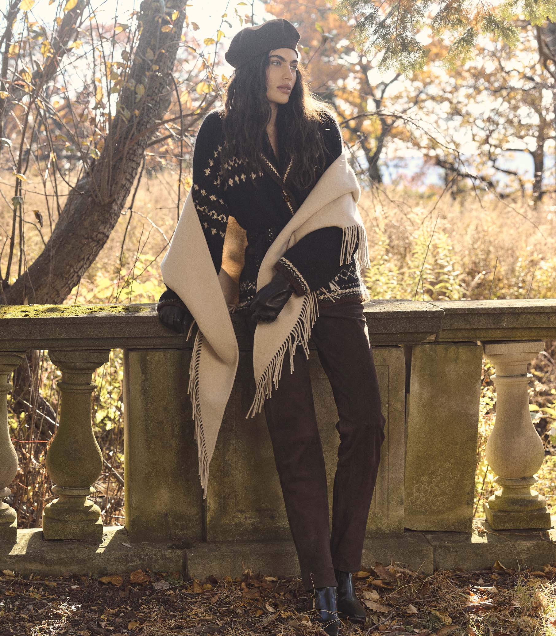 Woman in a black outfit with a cream shawl standing on a stone ledge in a natural setting.