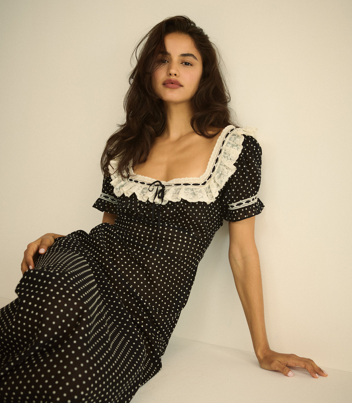 A woman wearing a black short-sleeve dress with white polka dots, a white lace-trimmed square neckline, and a small black bow at the center, posing against a plain off-white background. 1
