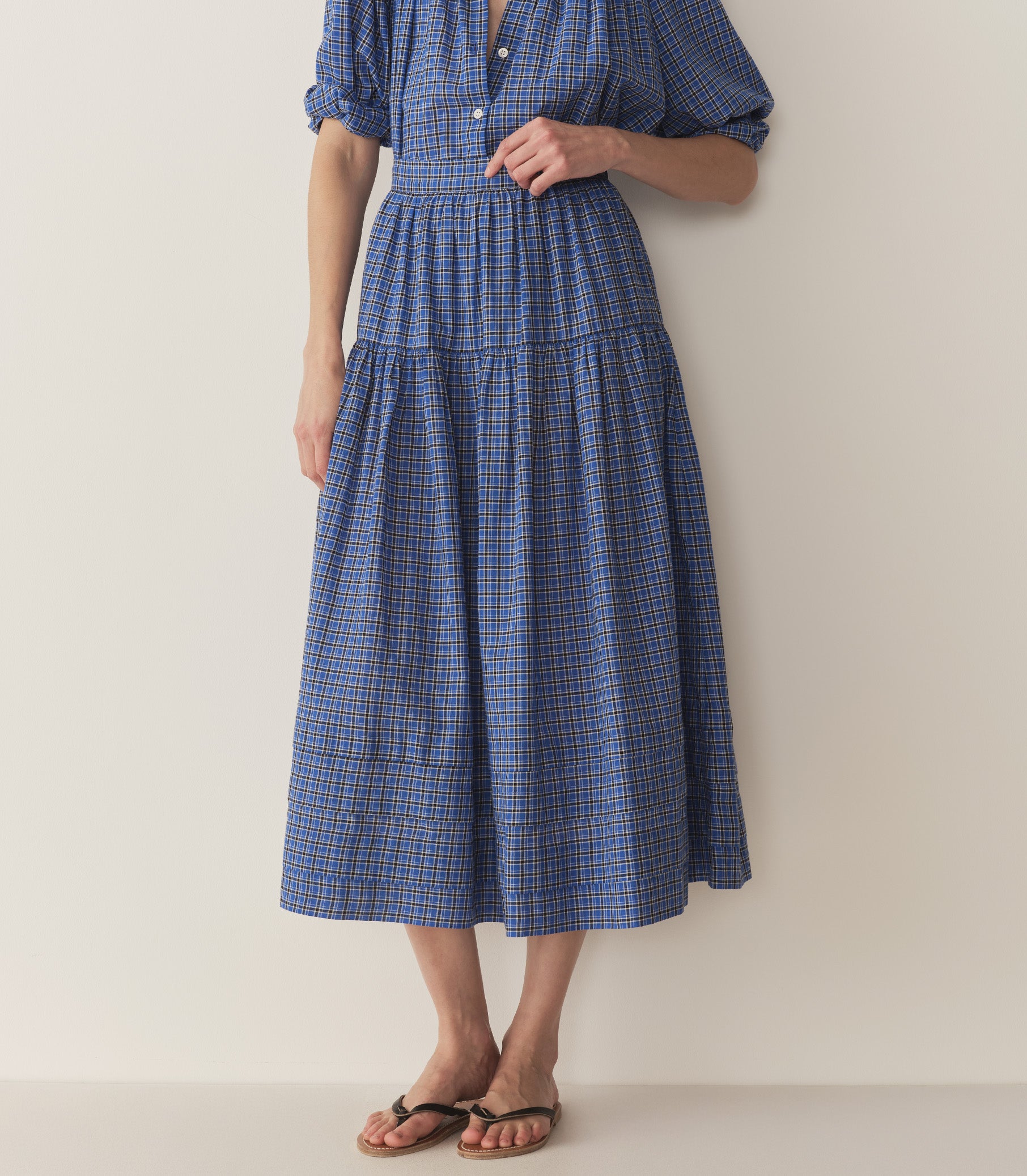 Bruna wears a blue and black checkered top and skirt with black flip flops.