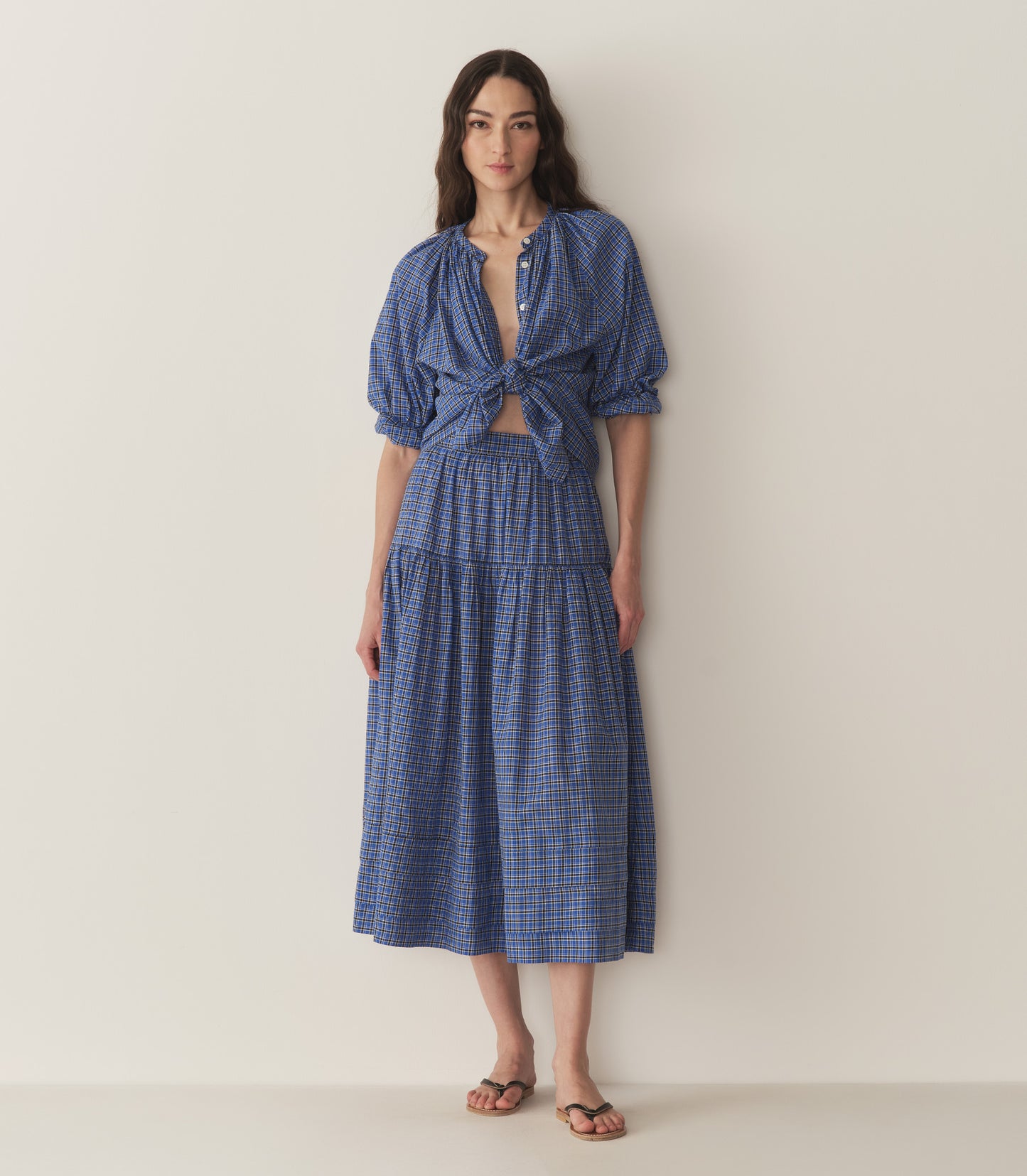 Bruna wears a blue and black checkered top and skirt with black flip flops.