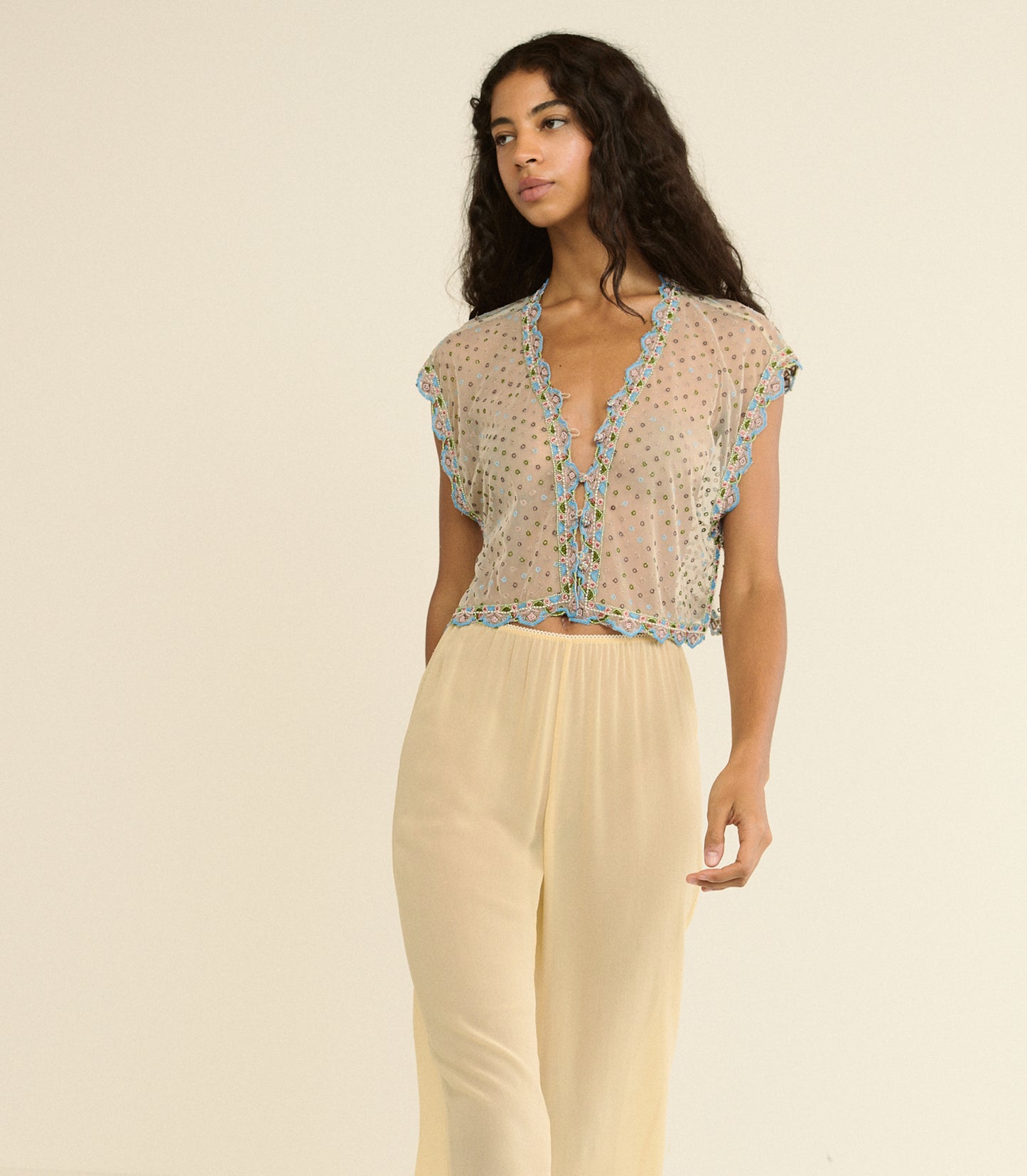 Woman wearing a sheer top with floral embellishments and light-colored pants on a beige background
