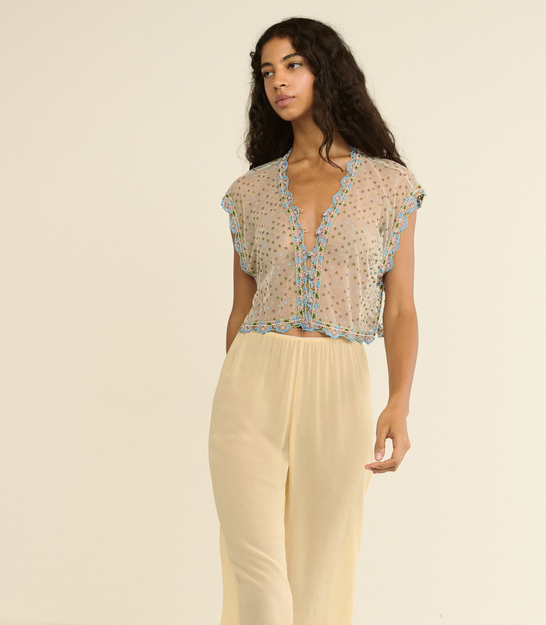 Woman wearing a sheer top with floral embellishments and light-colored pants on a beige background 1
