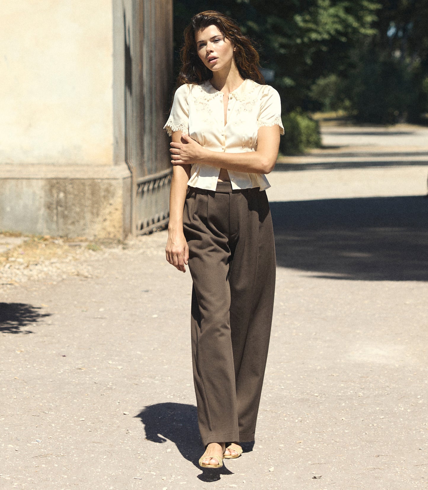 A woman stands outdoors on a sunlit paved path wearing a cream short-sleeve blouse with a ruffled collar and dark brown wide-leg trousers.