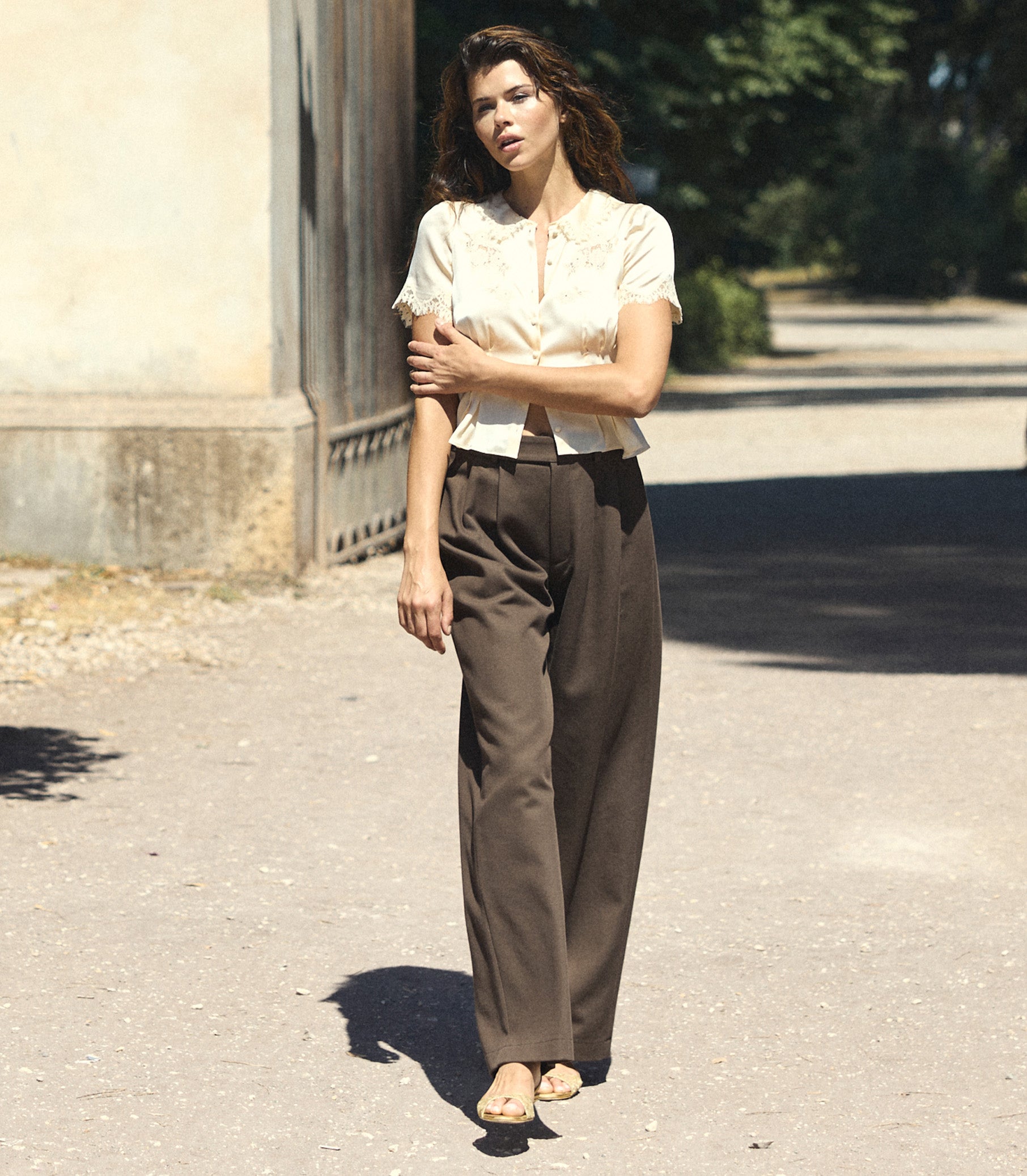 A woman stands outdoors on a sunlit paved path wearing a cream short-sleeve blouse with a ruffled collar and dark brown wide-leg trousers. 1
