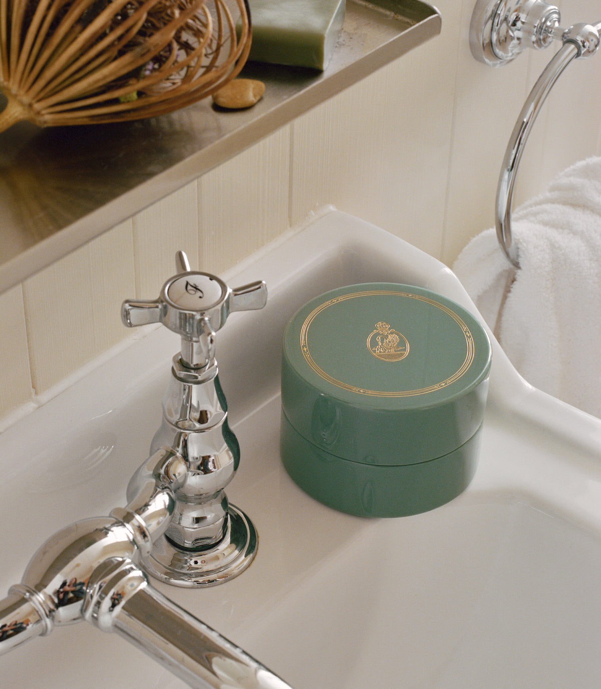 A round green cosmetic jar with a gold emblem on the lid, resting on a white bathroom sink next to a chrome faucet. 1
