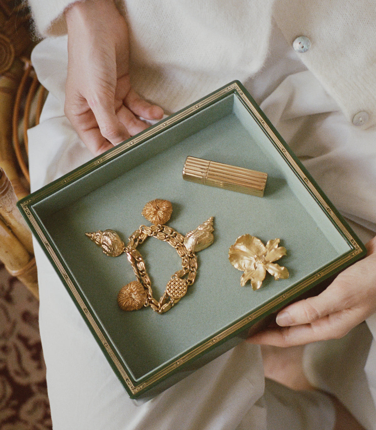 A person in a cream-colored coat holds an open dark green jewelry box with gold trim, displaying three gold vintage-style accessories: a textured chain necklace, a small gold cylindrical piece, and a gold flower-shaped accessory. 1
