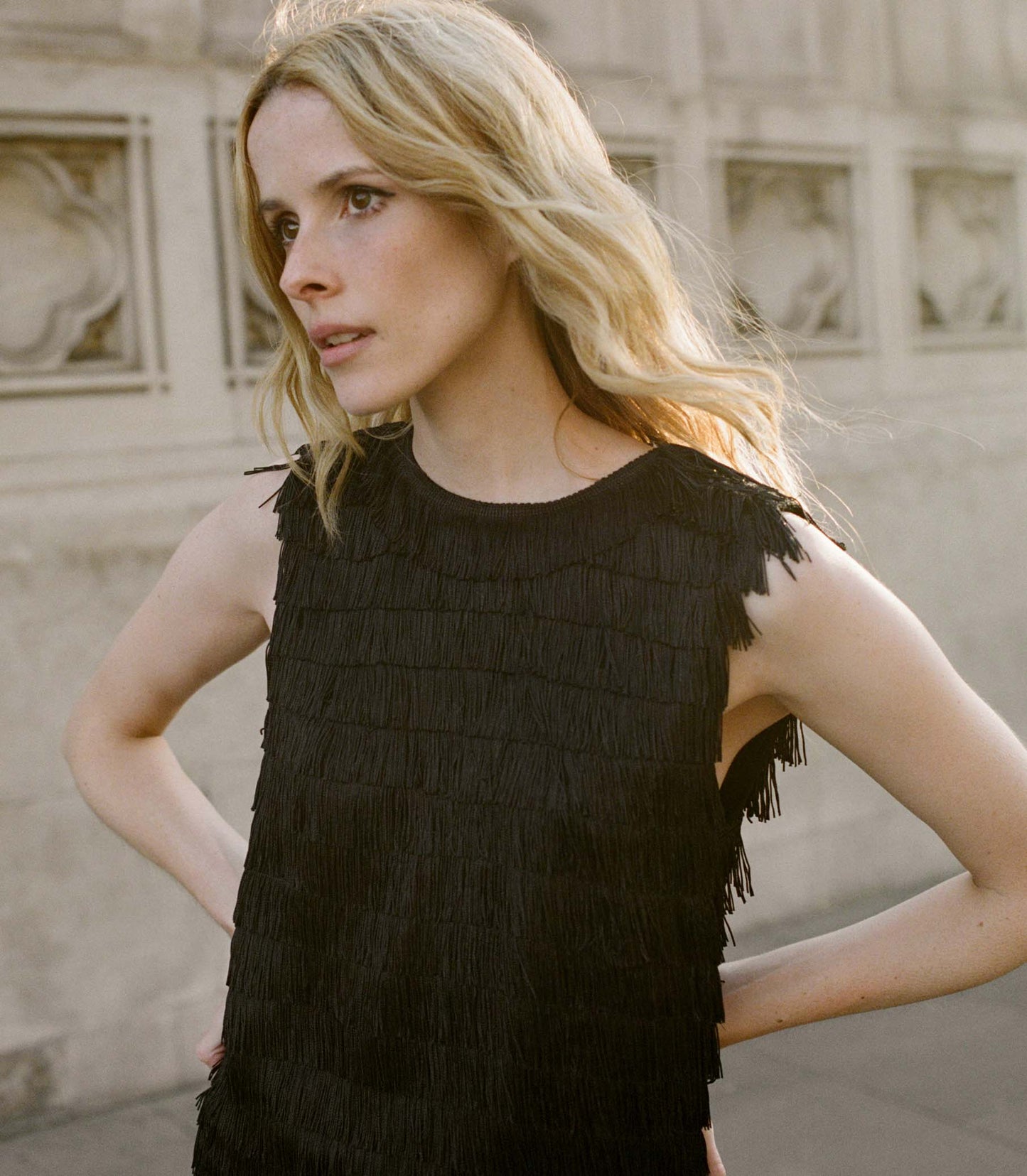 Woman wearing a black fringed top standing in front of a stone wall.