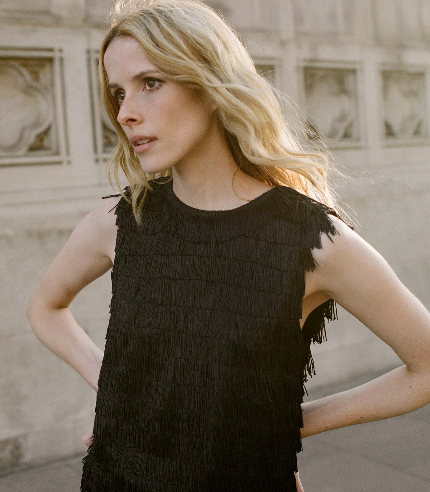 Woman wearing a black fringed top standing in front of a stone wall. 1