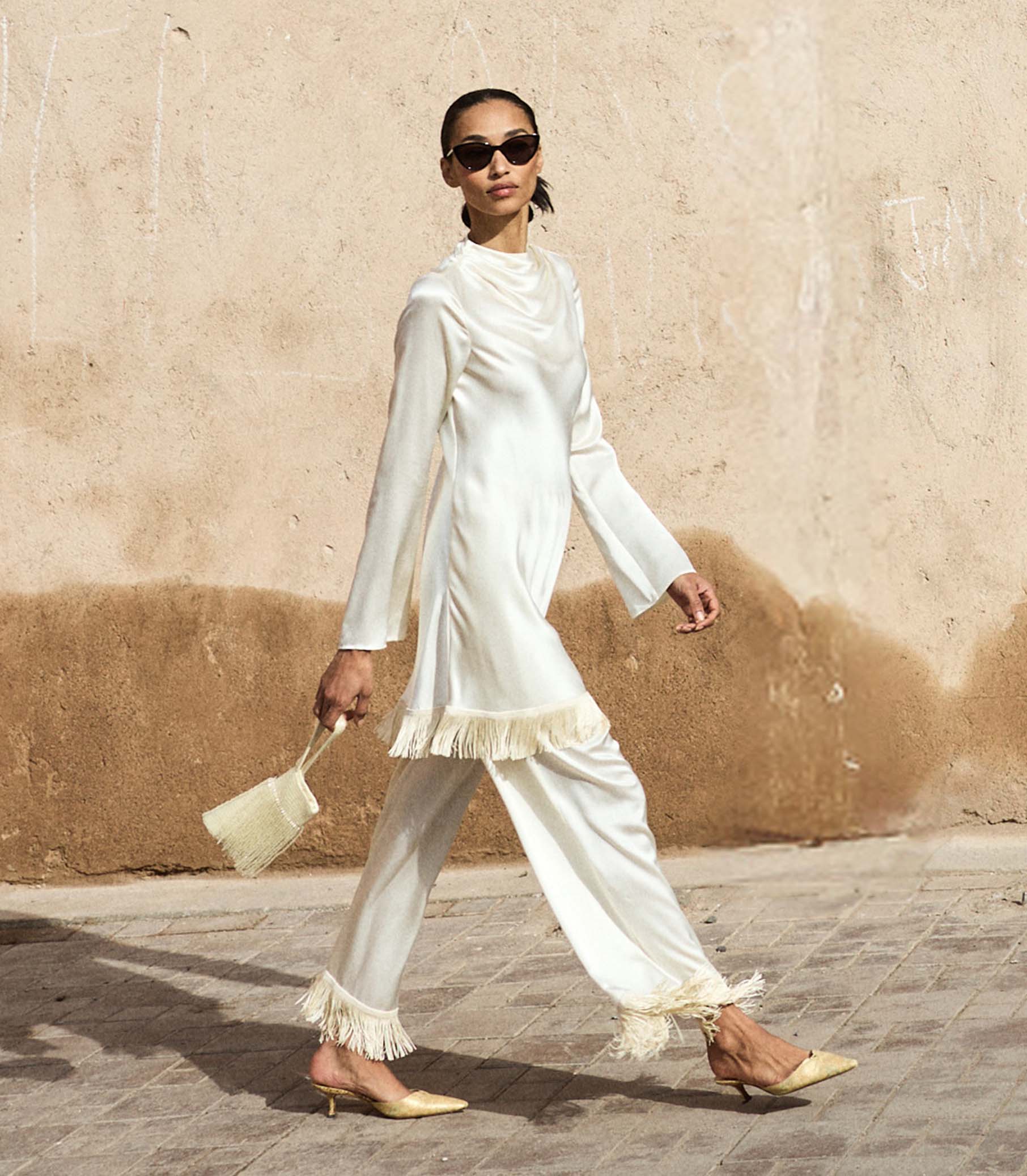 Woman in a white fringe dress walking on a street with a beige wall in the background