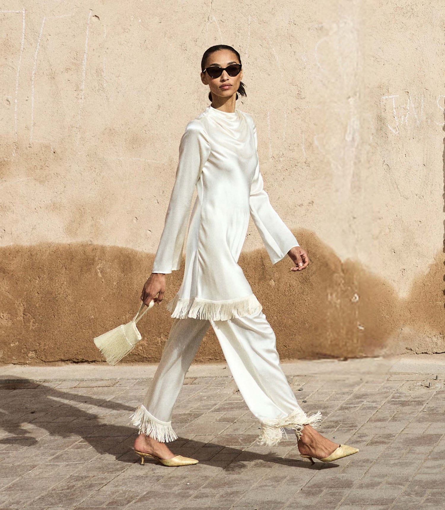 Woman in a white fringe dress and matching pants walking on a street with a beige wall in the background