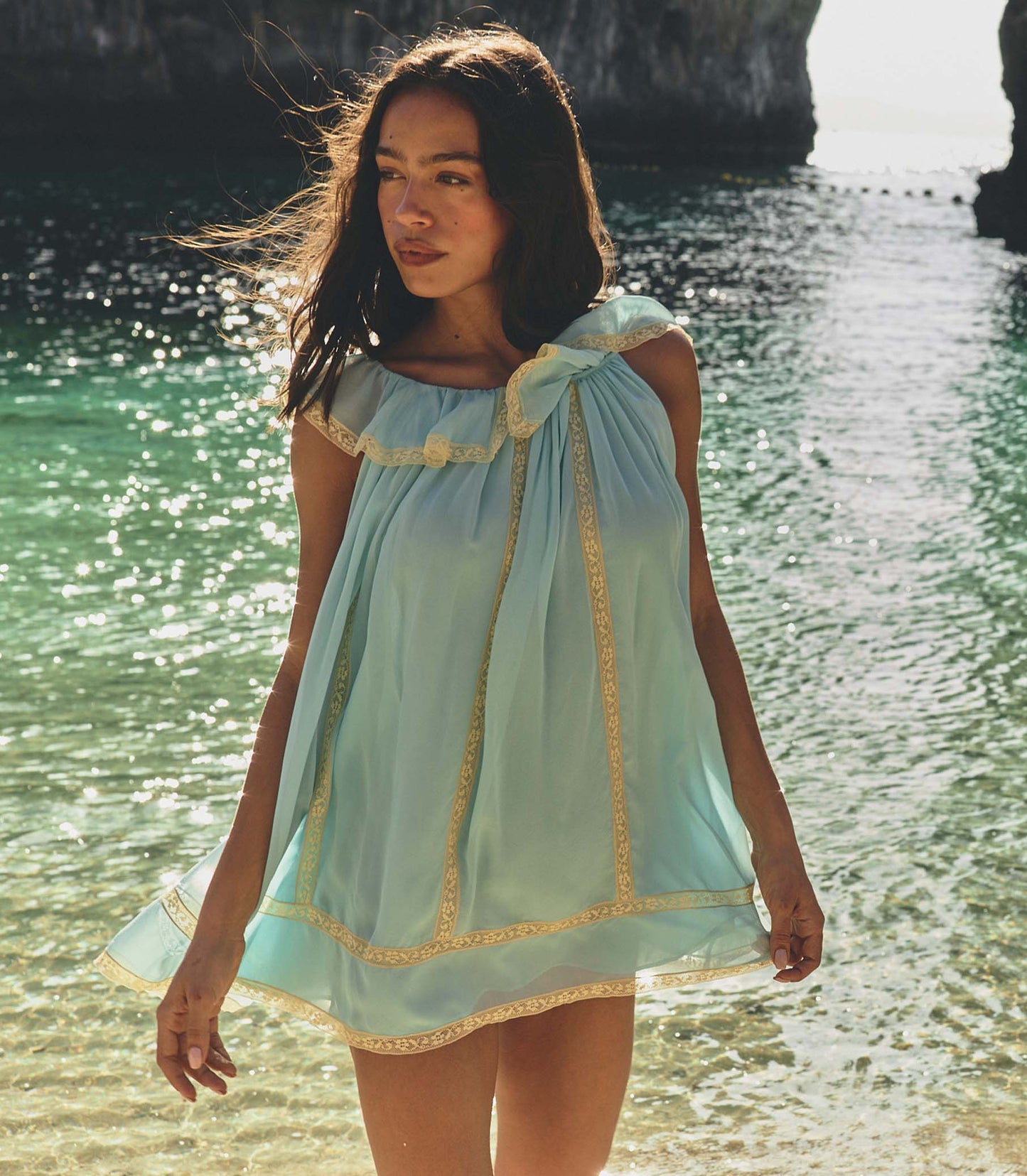 Woman in a light blue dress standing by a body of water with sunlight filtering through.