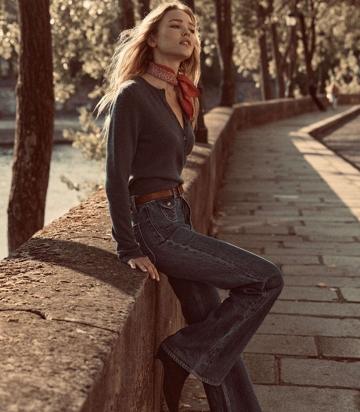 A woman wearing a dark charcoal knit sweater, blue flared jeans, and a red patterned neck scarf sits on a stone bridge in a park with trees and a paved walkway.