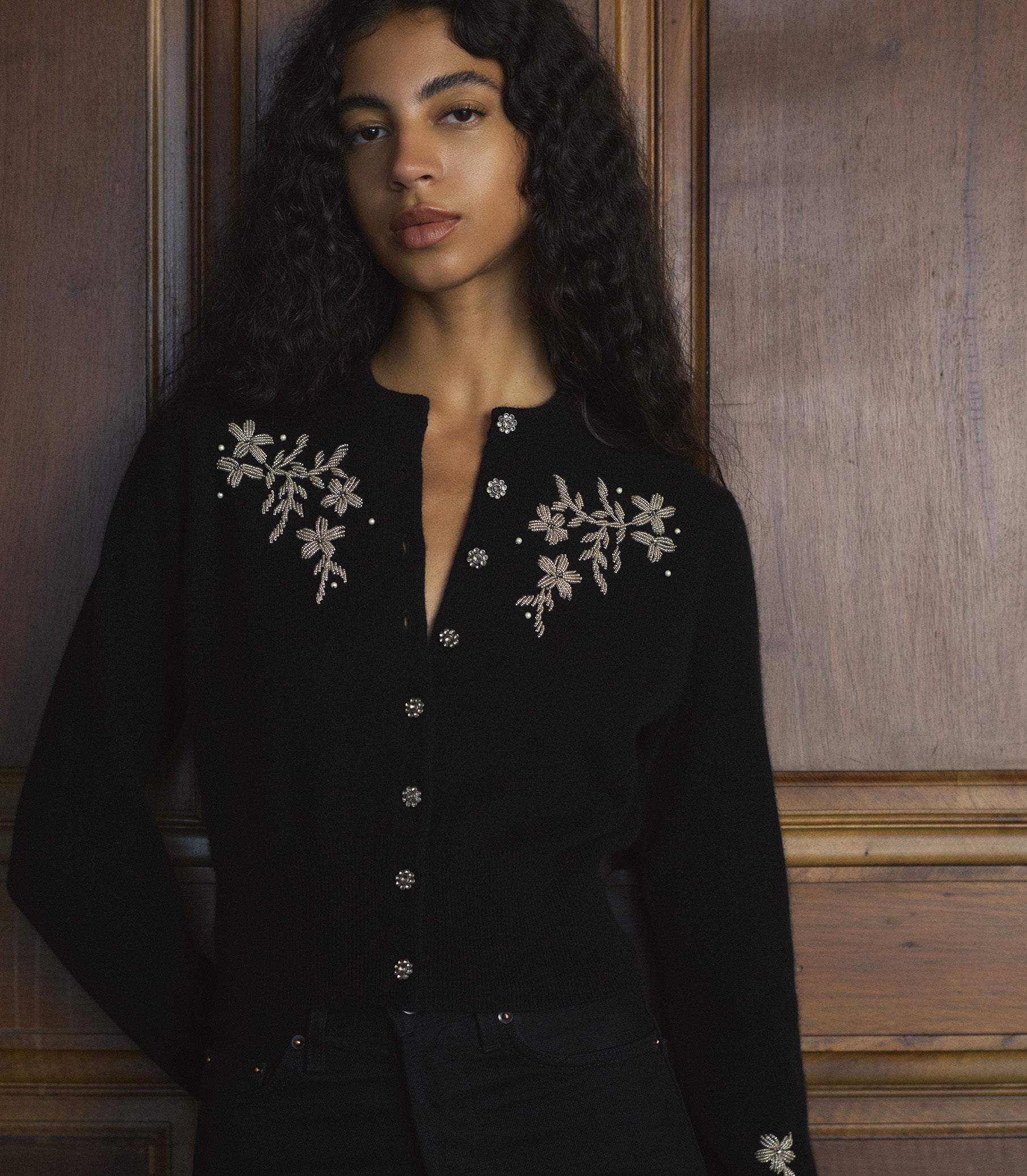 Woman wearing a black cardigan with floral embroidery against a wooden paneled wall. 1