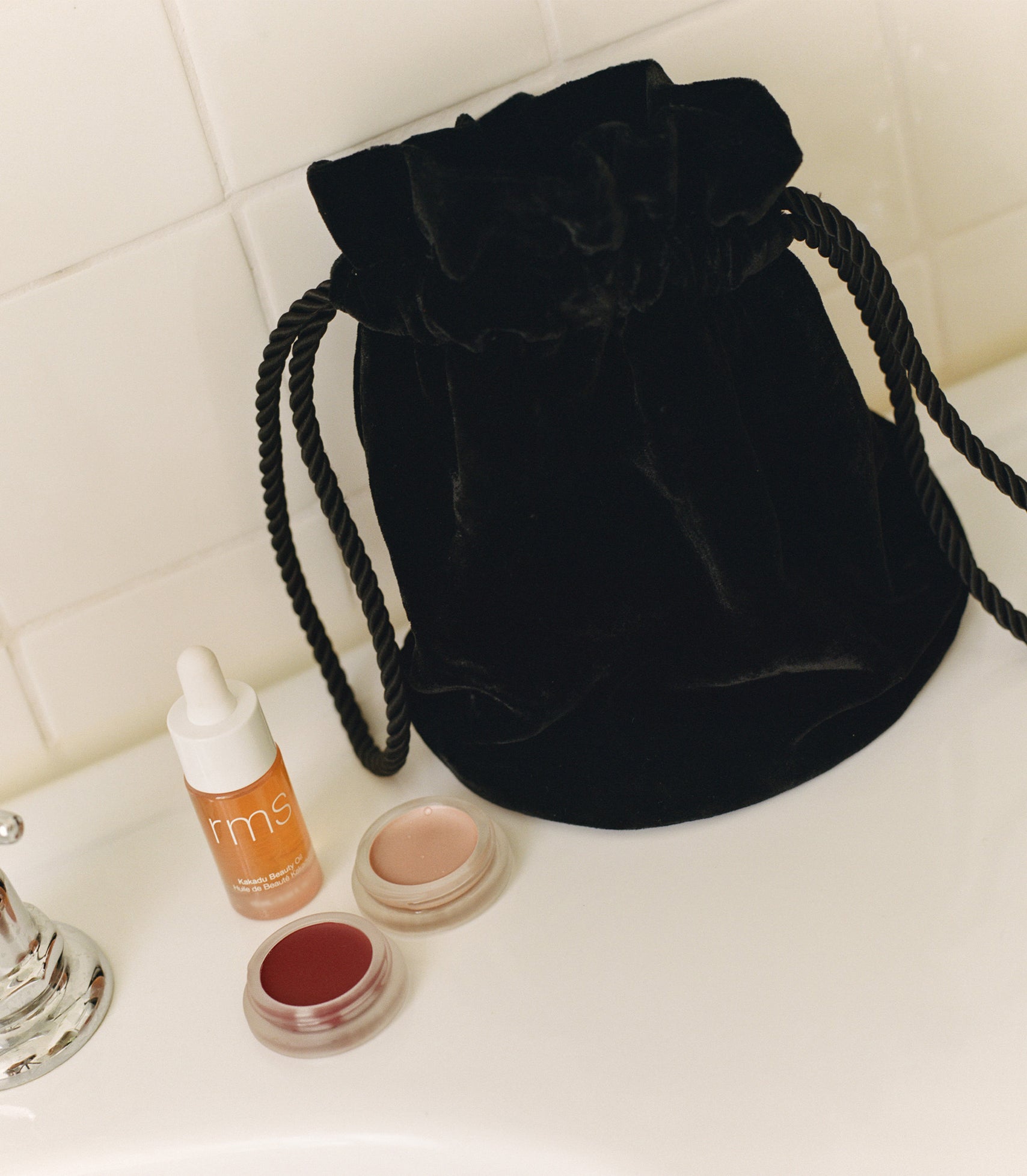 A black velvet pochette on a counter next to a lip stain, cheek luminizer, and skincare oil.