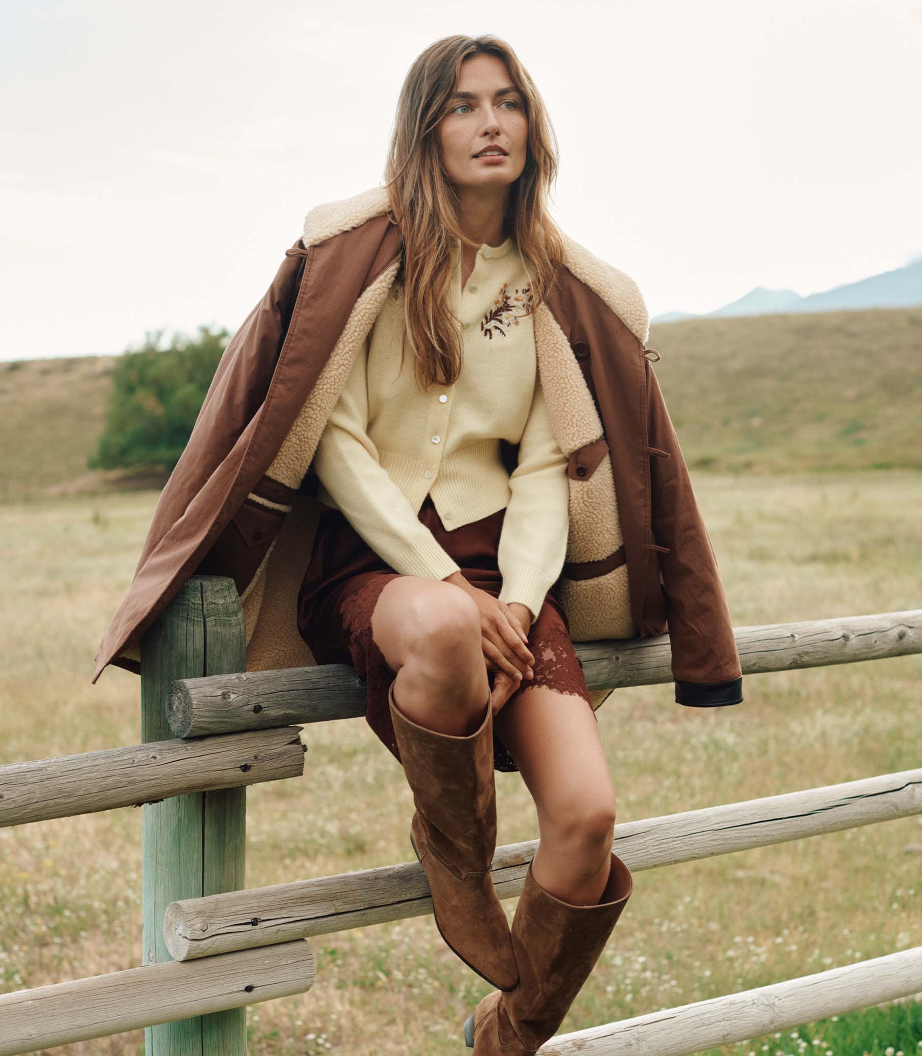 Woman sitting on a wooden fence in a field wearing a brown coat and knee-high boots. 2