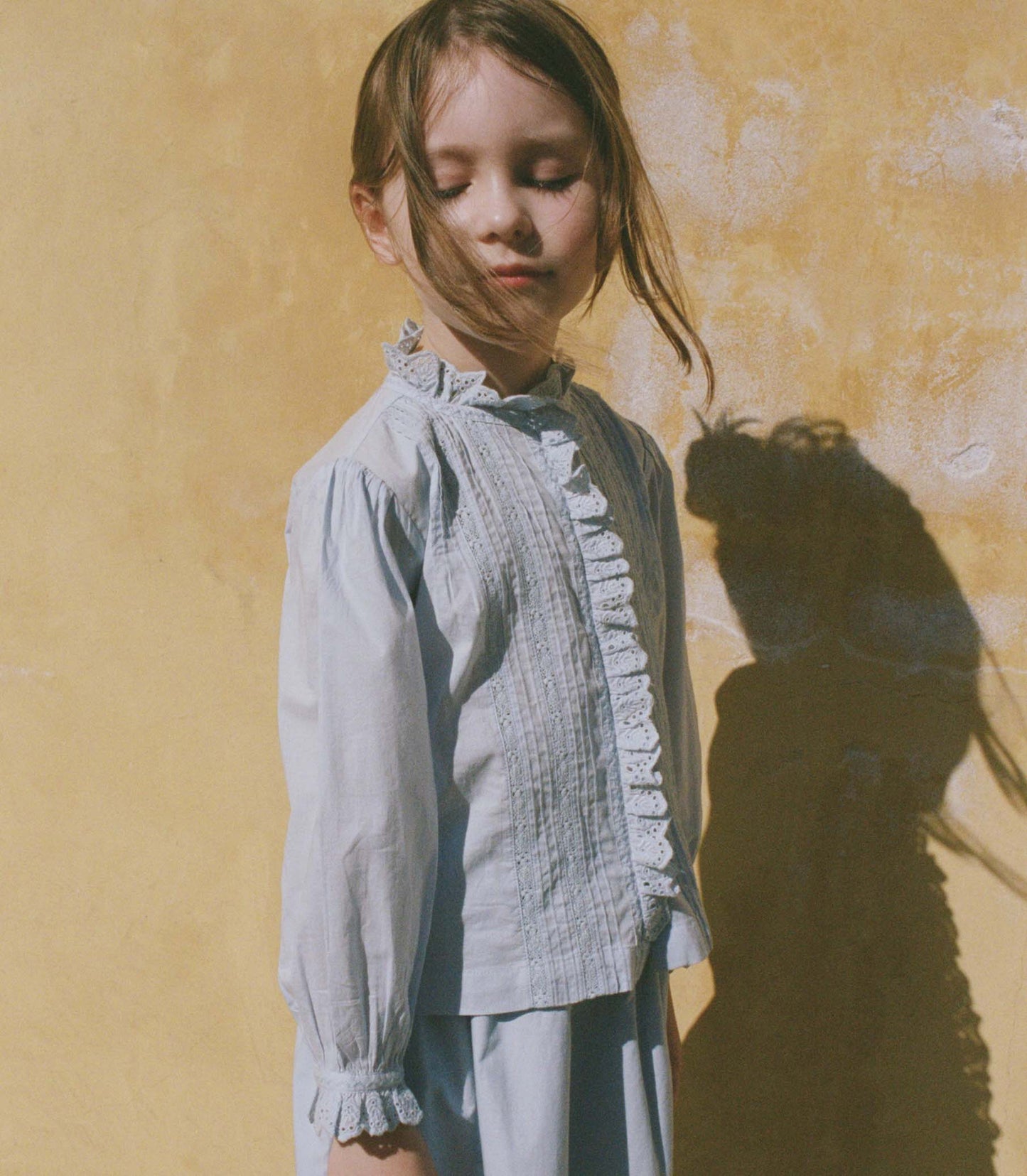Young girl in a light blue blouse standing against a textured yellow wall with a soft focus.