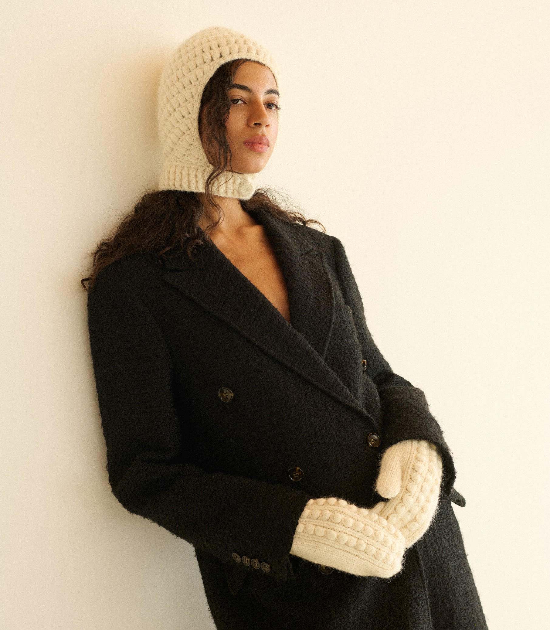 Woman wearing a black coat, cream knitted bonnet, and gloves against a beige background 1