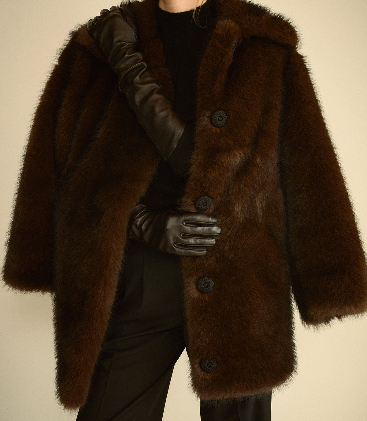 A person wearing a dark brown plush fur coat with large black buttons, black gloves, and a black top, photographed against a plain light beige background.