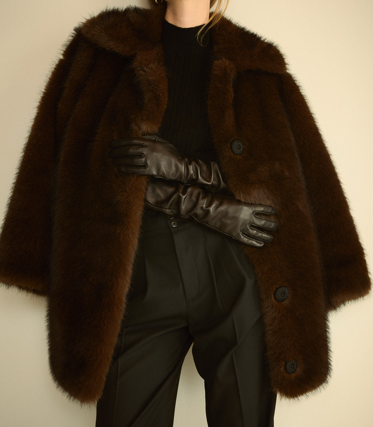A person wearing a dark brown shaggy faux-fur coat over a black ribbed top and black pants, with long black gloves crossed at the waist, against a plain light beige background.