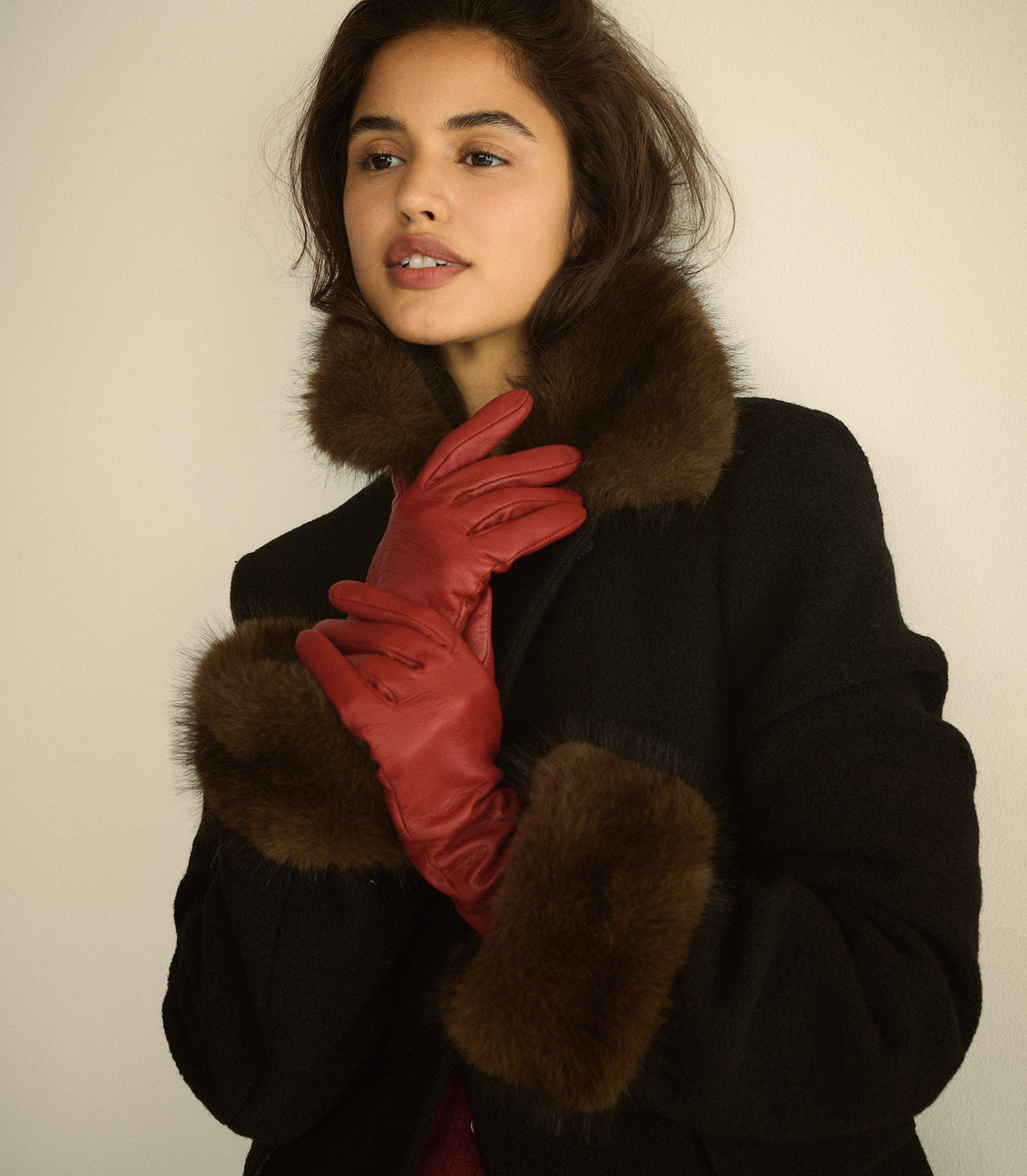 A woman wearing a black coat with brown fur trim and red leather gloves, posed against a plain light beige background.