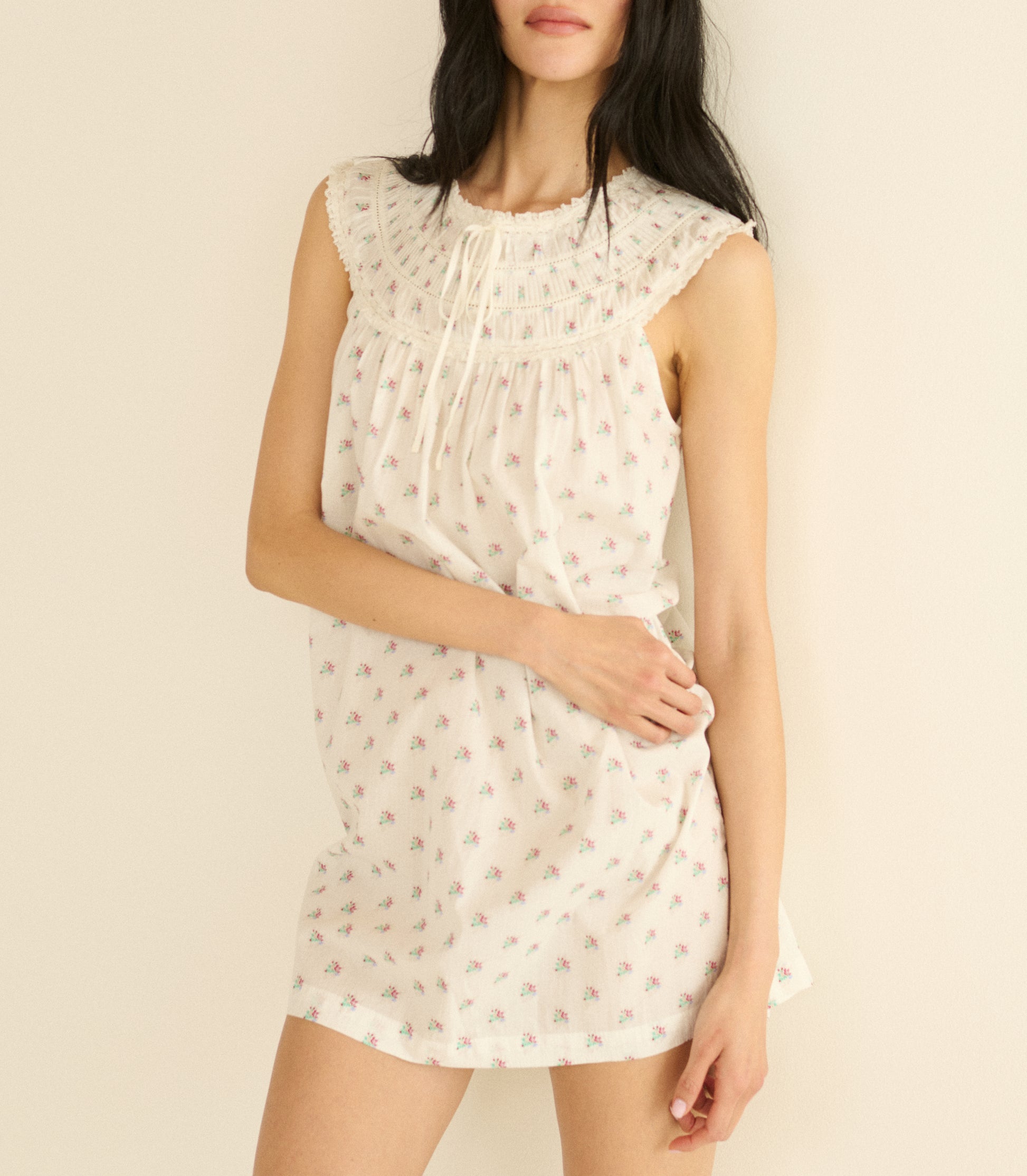 Woman wearing a white sleeveless dress with a subtle floral pattern on a plain background 1
