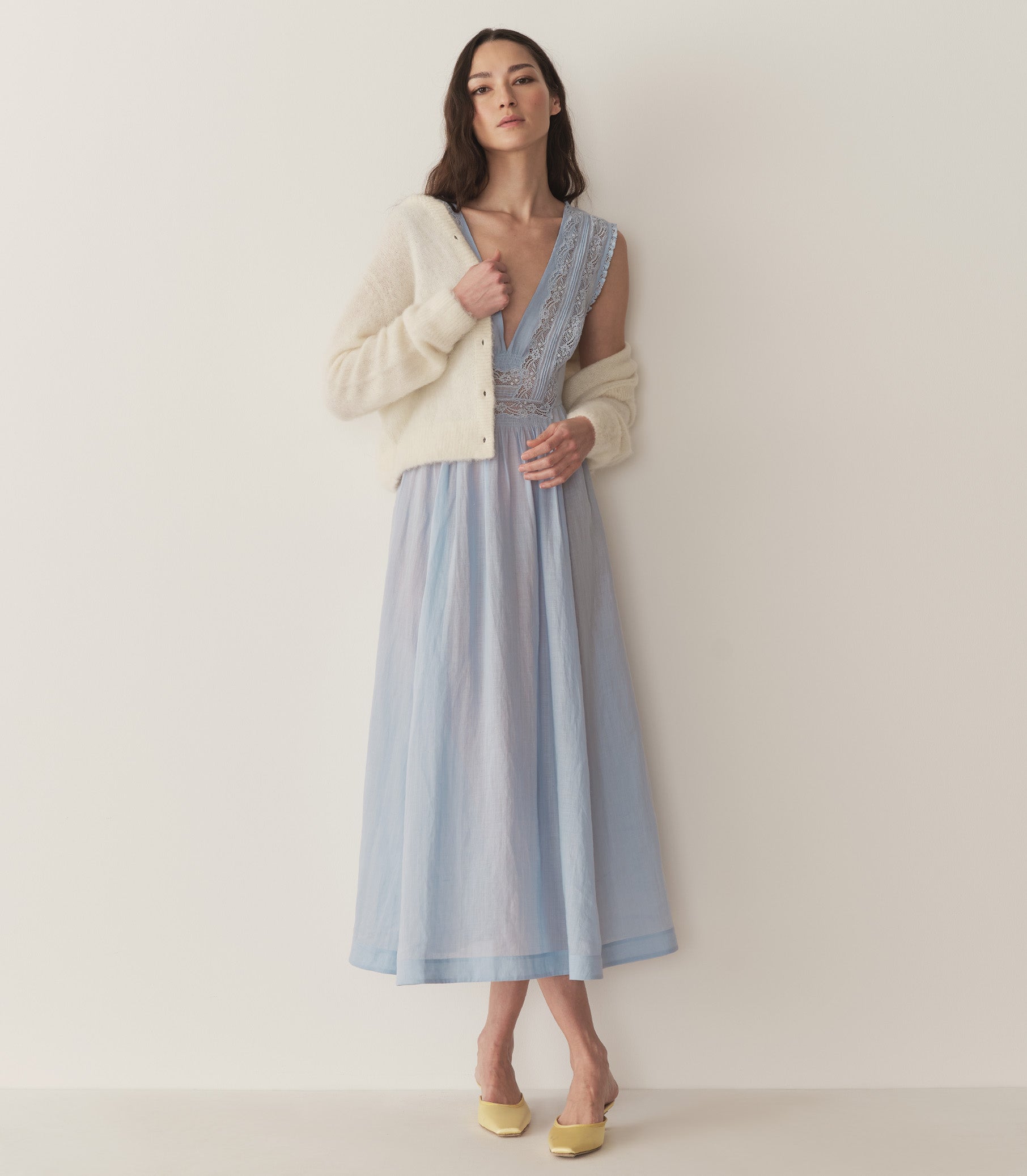 Bruna is wearing a long light blue sleeveless dress with lace details and light yellow kitten heels. 2