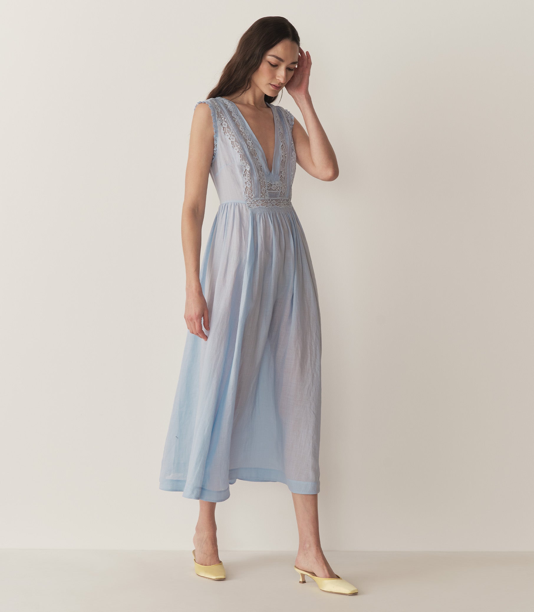 Bruna is wearing a long light blue sleeveless dress with lace details and light yellow kitten heels. 1