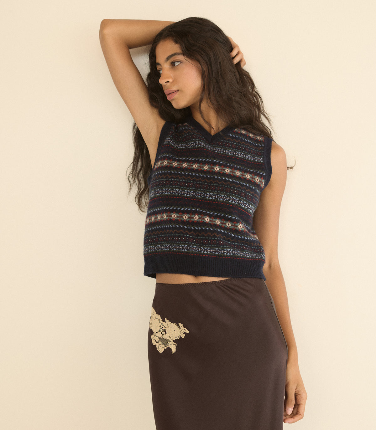 A woman models a sleeveless cropped knit top with horizontal geometric patterns in navy, beige, and white, paired with a dark brown skirt against a plain cream background.