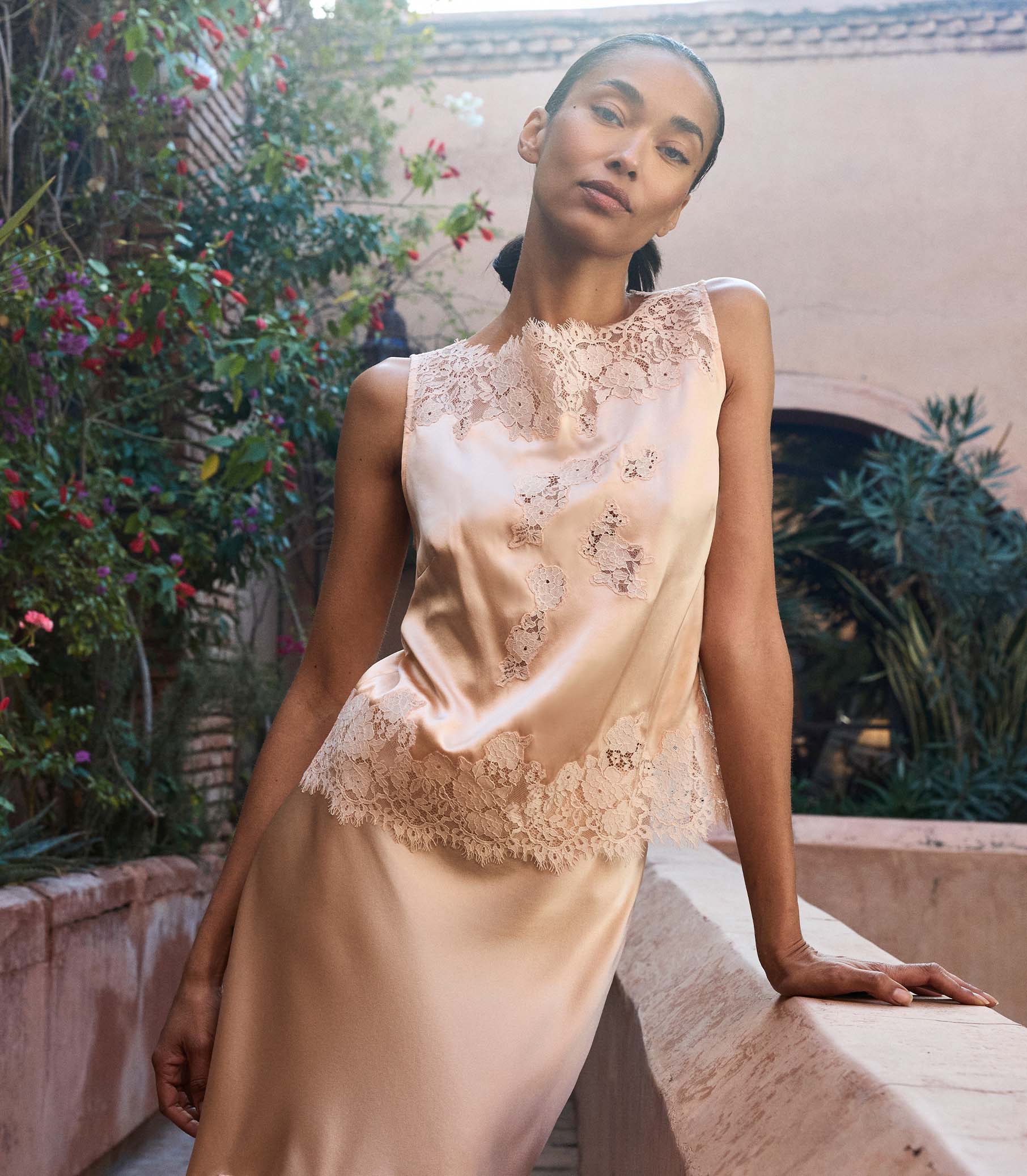 Woman wearing a peach lace blouse with a matching skirt sitting outdoors with greenery in the background