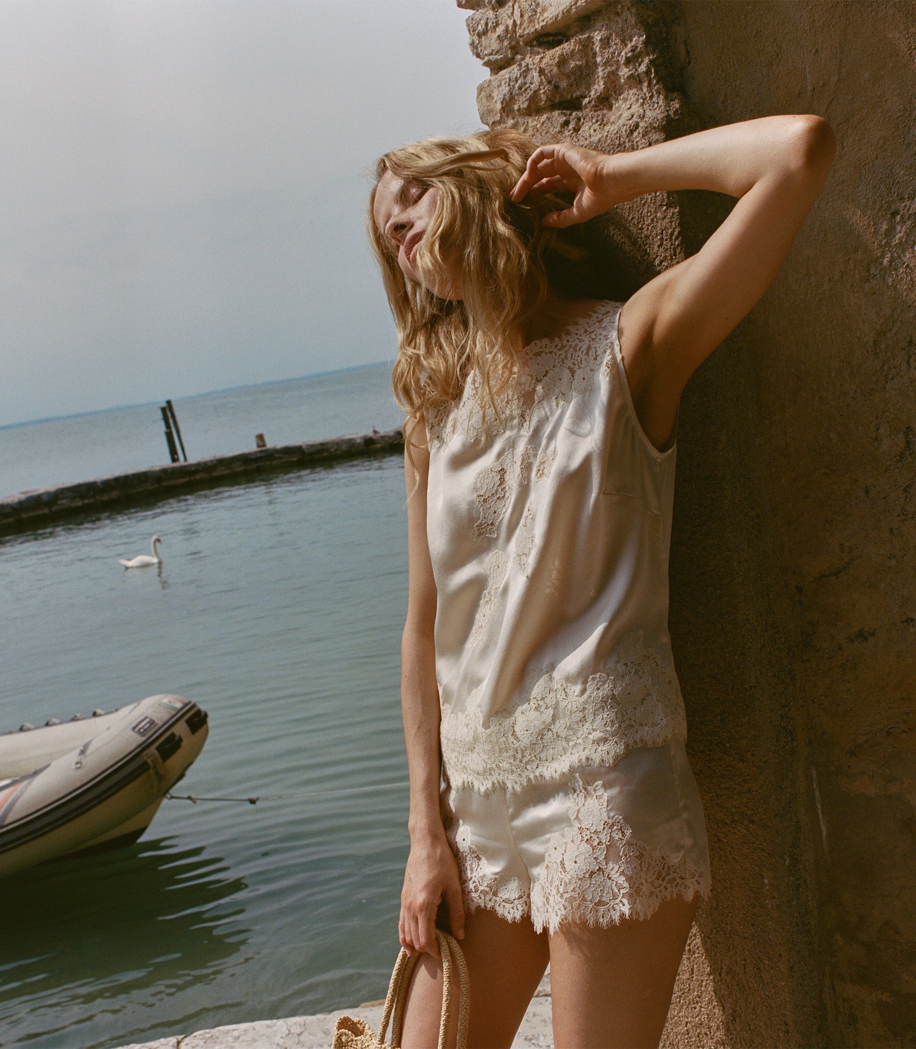 Woman in a white lace outfit leaning against a stone wall by a body of water with a swan and boat. 1