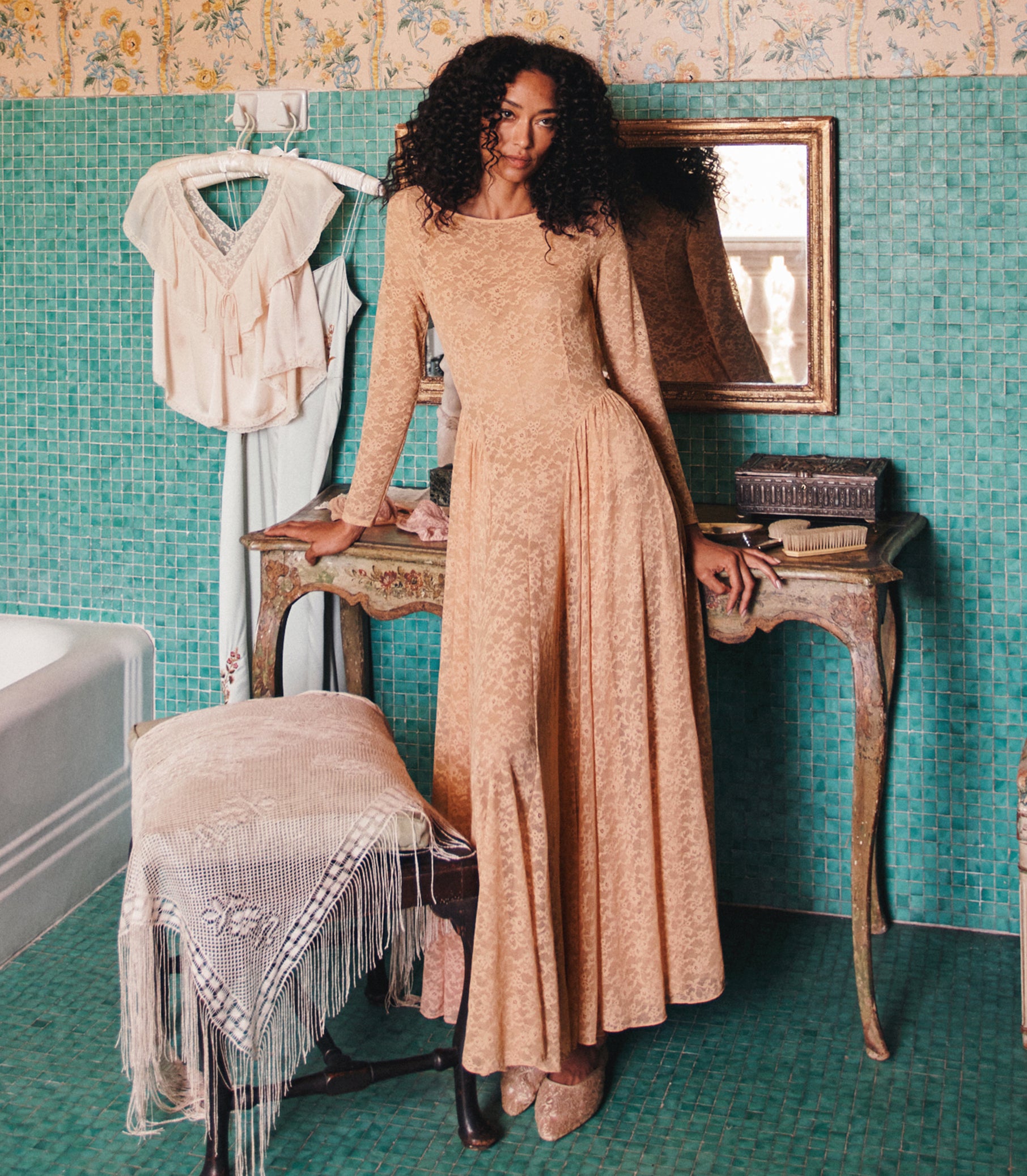 A woman with curly dark hair models a long-sleeved beige lace maxi dress in a vintage-style room with teal mosaic tile walls, leaning on a wooden table. 1
