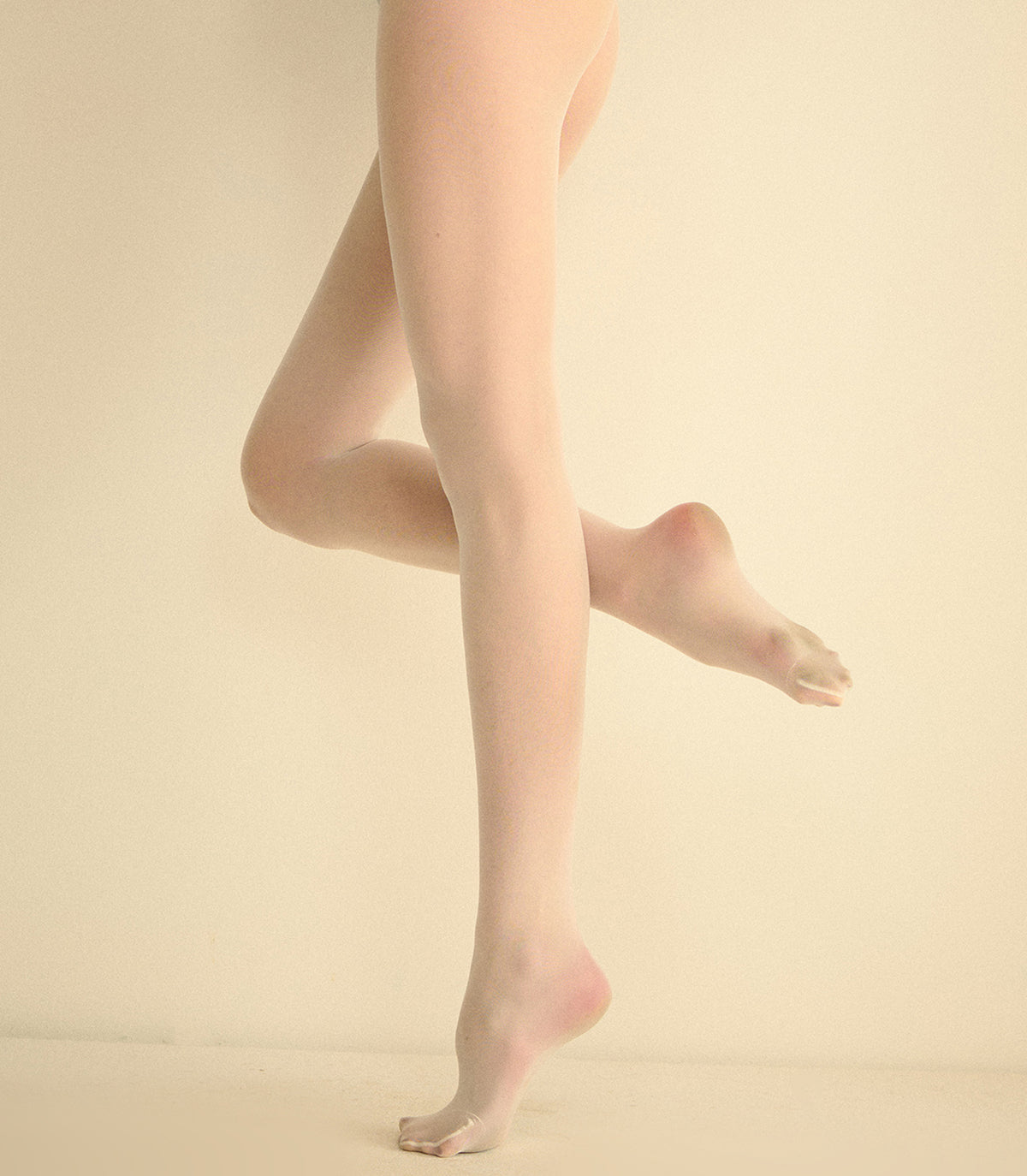 A close-up of a person's legs and feet wearing sheer nude tights, posed against a plain warm beige studio background.