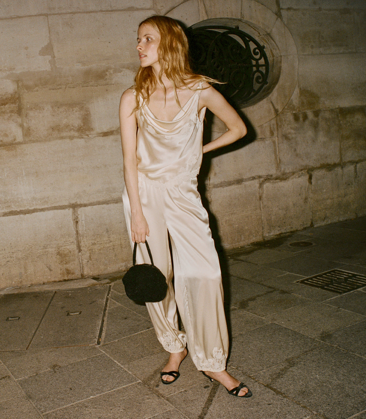 A woman stands on a stone sidewalk against a stone wall, wearing a satin-like beige jumpsuit with draped straps and loose harem-style legs. She holds a small black round handbag and wears black flat sandals. 1
