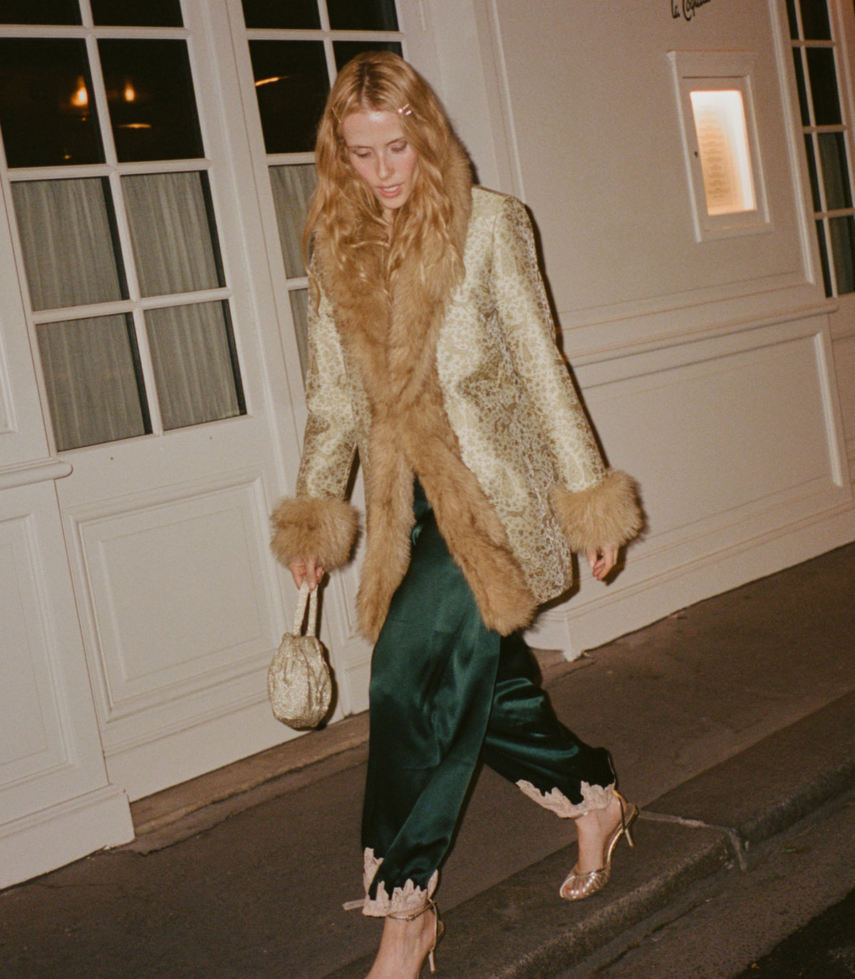 A woman walking on a sidewalk at night wearing a beige and brown animal-print faux fur coat with a large tan fur collar, dark green satin pants, and holding a small beige fur handbag.