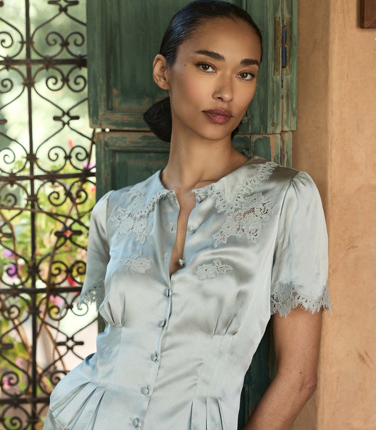 Woman wearing a light blue silk blouse with lace details standing in front of a decorative metal gate.