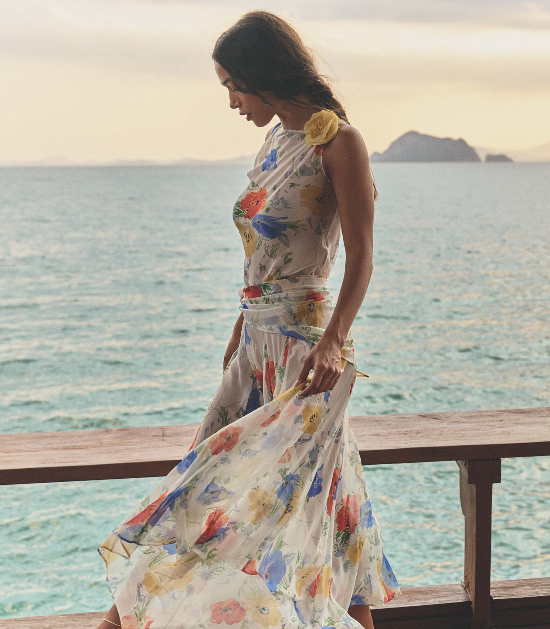 Woman in a white floral dress standing on a wooden platform by the ocean with mountains in the background. 1
