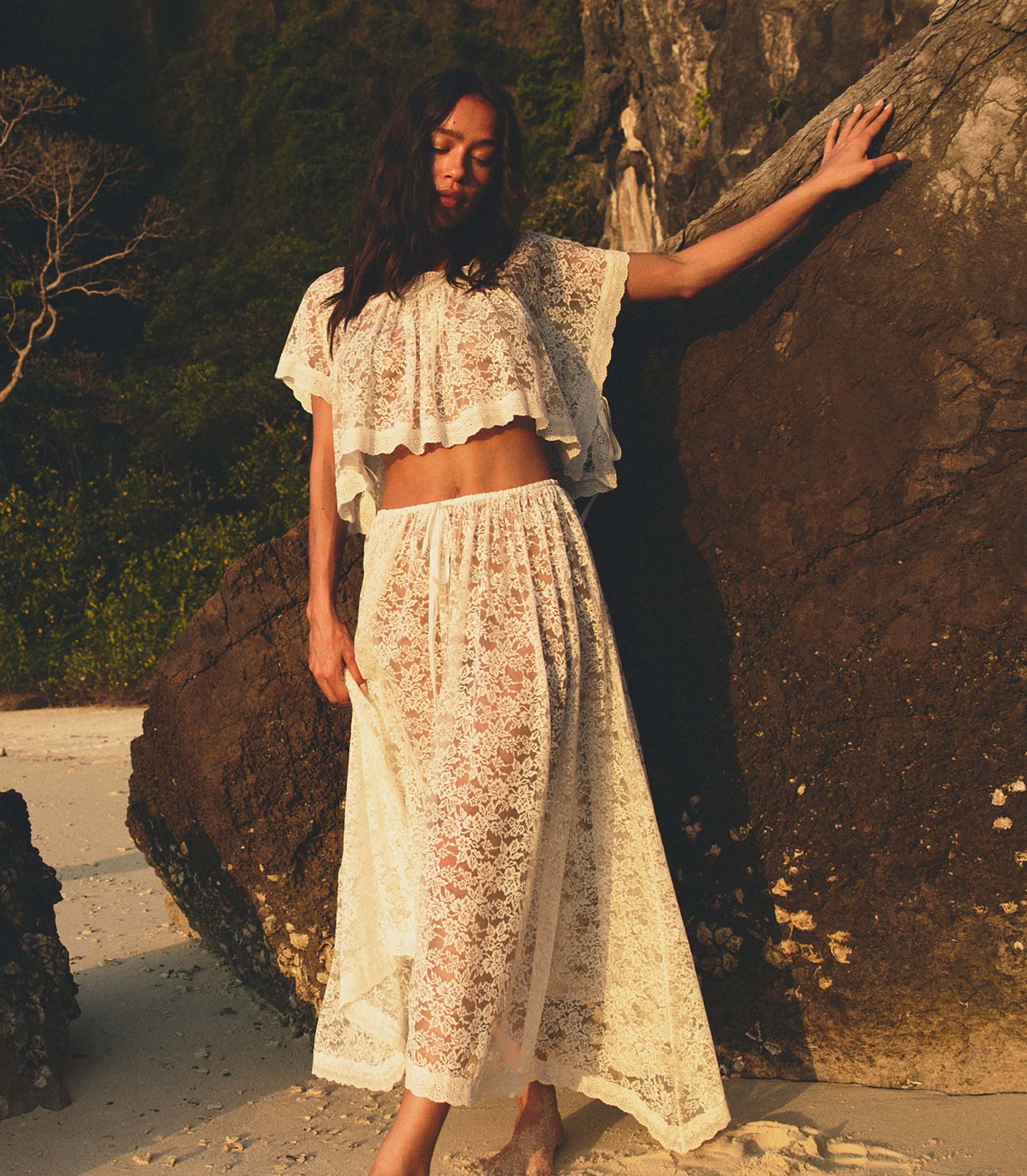 Woman in a white lace top and matching skirt standing against a rock with a natural background